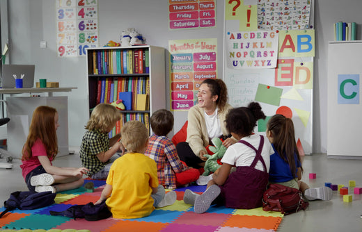 A group of young children sitting on a colorful foam mat in a preschool classroom, attentively listening to their teacher who is holding a plush toy. The classroom features educational posters, books, and learning materials.