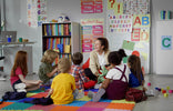 A group of young children sitting on a colorful foam mat in a preschool classroom, attentively listening to their teacher who is holding a plush toy. The classroom features educational posters, books, and learning materials.