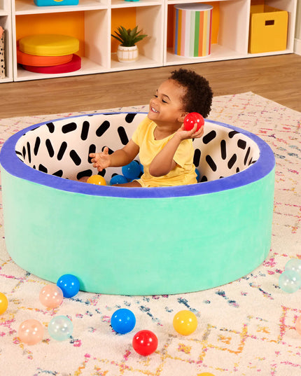 Child playing with colorful balls inside a green playpen with black and white pattern on a patterned rug.
