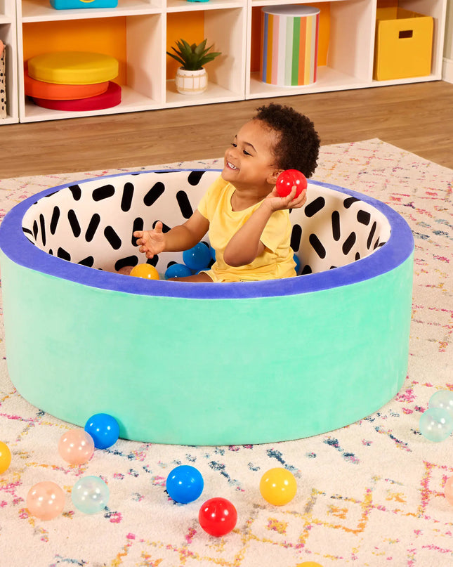 Child playing with colorful balls inside a green playpen with black and white pattern on a patterned rug.