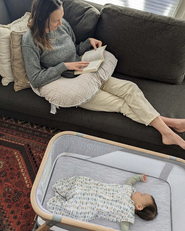 Woman reading a book on a couch next to a baby in a crib.