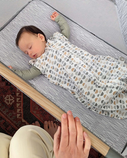 Baby sleeping in a patterned swaddle on a changing table with a person's hand visible.