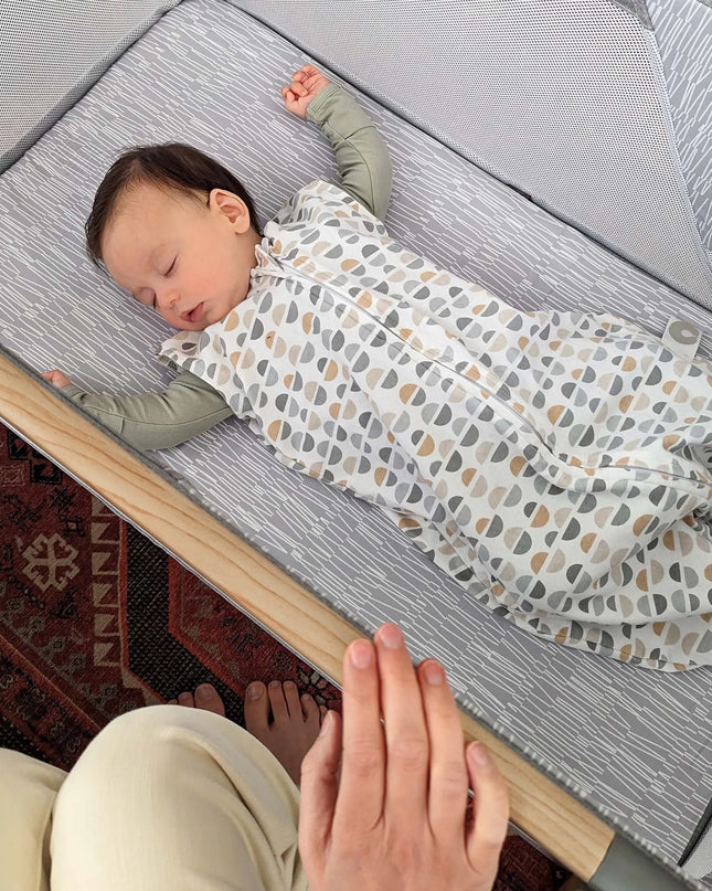 Baby sleeping in a patterned swaddle on a changing table with a person's hand visible.