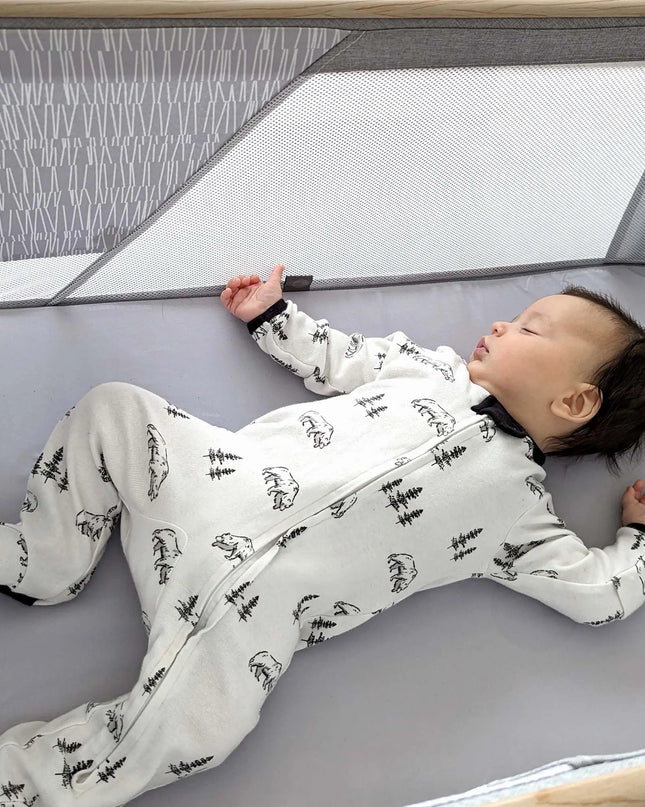 Baby lying on a gray playpen wearing a white onesie with black patterns.