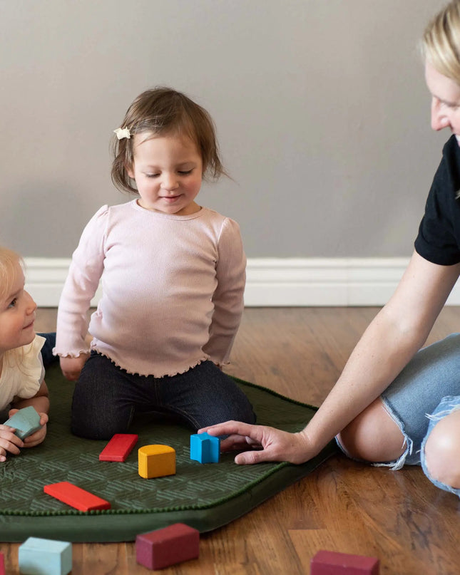 Two children playing with colorful blocks on a green mat, assisted by an adult.