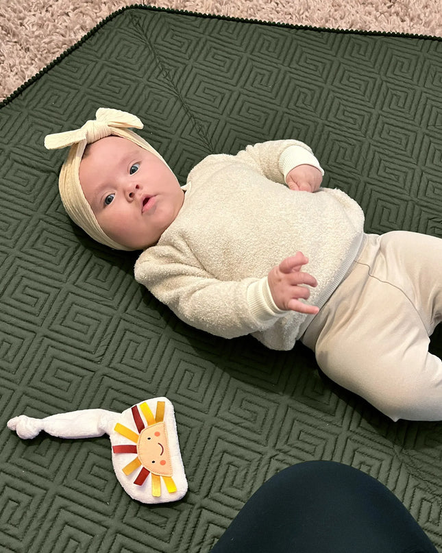 Baby lying on a green play mat with a toy, wearing a white outfit and headband.