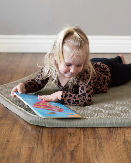 Child reading a book on a cushioned mat on a wooden floor.