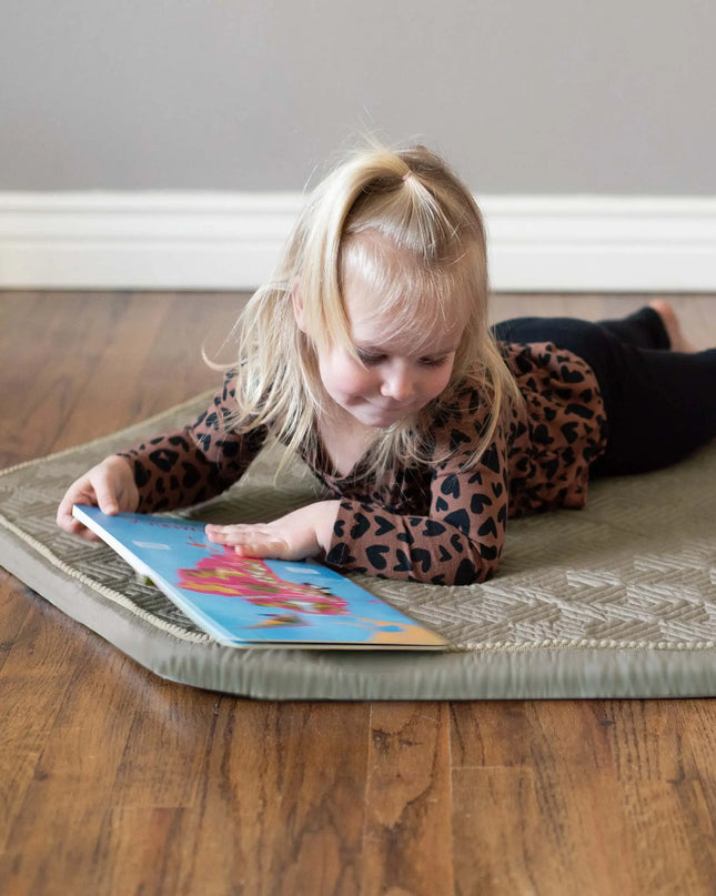 Child reading a book on a cushioned mat on a wooden floor.