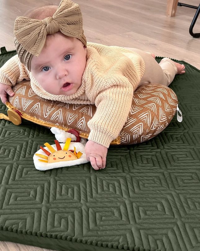 Baby sitting on a green mat with a toy, wearing a beige outfit and headband.