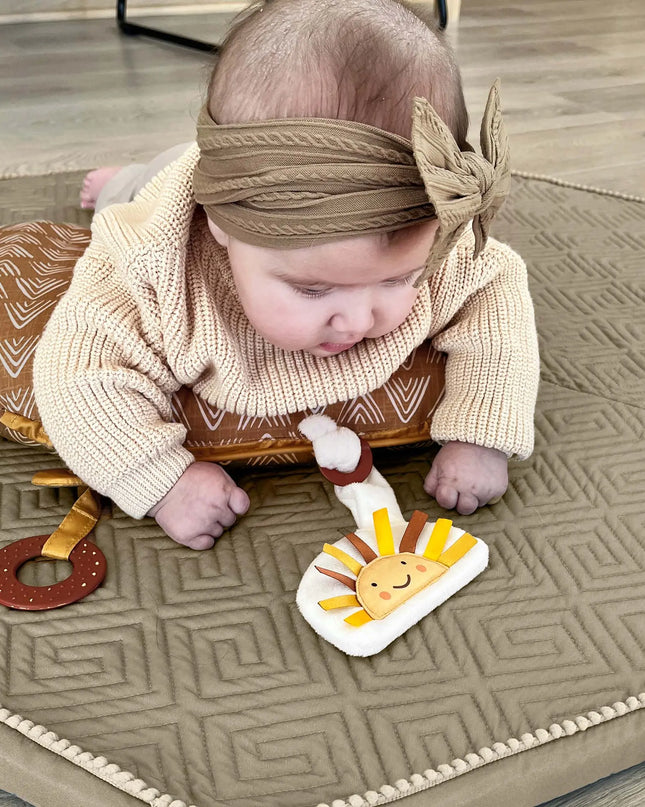 Baby crawling on a patterned mat with toys, wearing a headband.