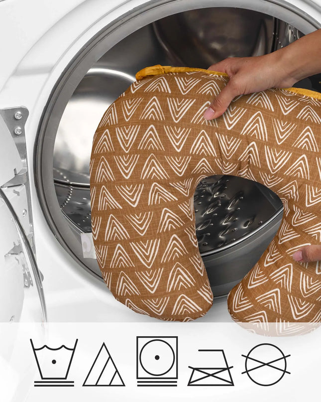 Patterned travel pillow held by a hand in front of an open washing machine with laundry symbols below.