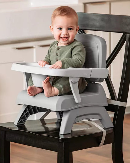 Baby sitting in a gray high chair attached to a wooden chair in a kitchen.