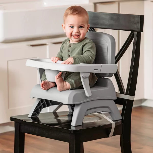 Baby sitting in a gray high chair attached to a wooden chair in a kitchen.