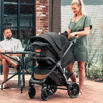 Woman pushing a stroller outdoors with a man sitting at a table in the background.