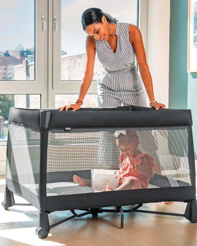 Woman and child in a travel crib in a bright room with large windows.