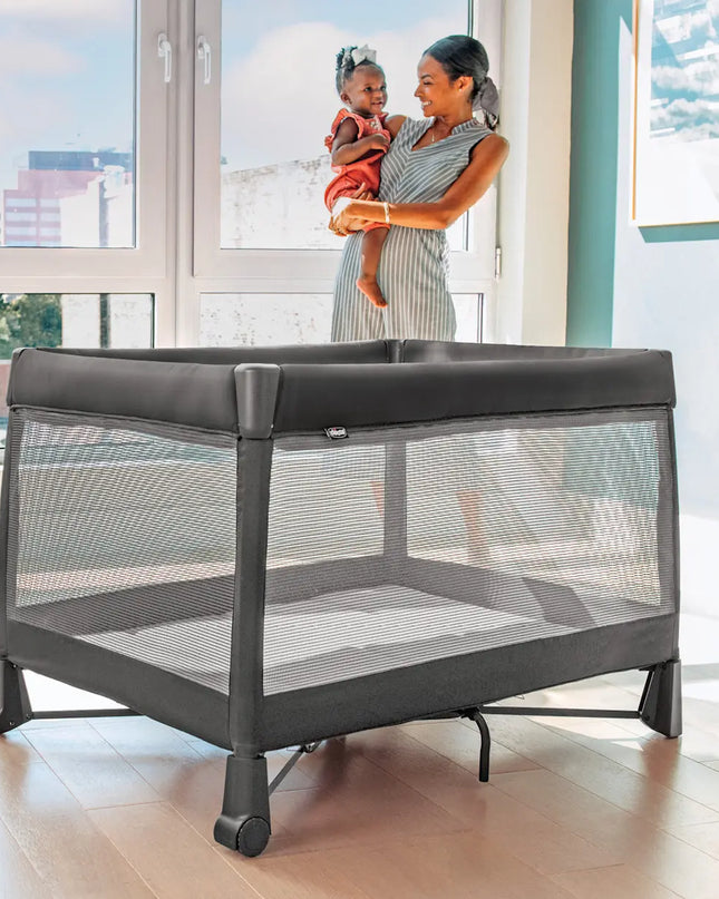 Woman holding a child next to a gray playpen in a room with large windows.