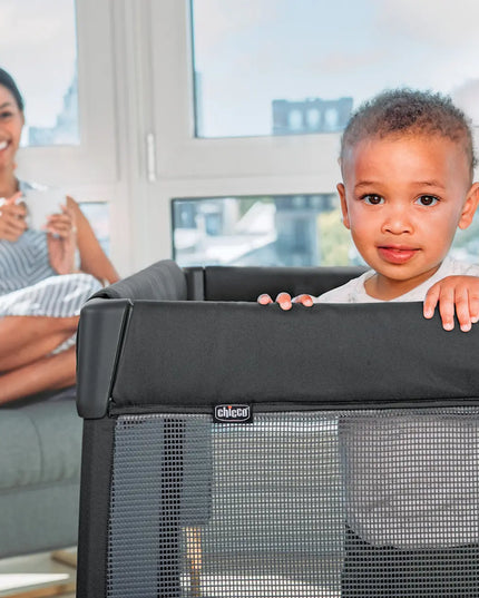 Child peeking over a black baby gate with a woman sitting on a couch in the background.