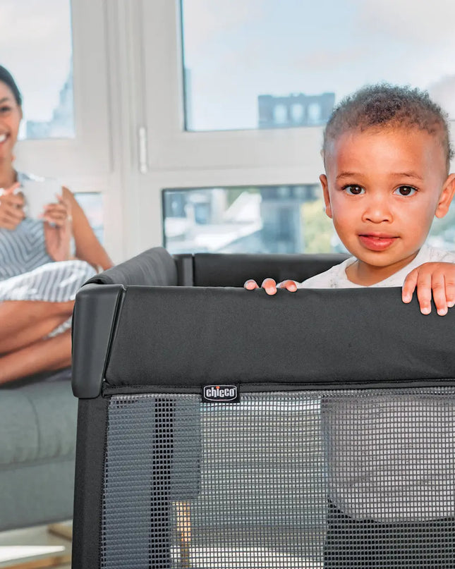 Child peeking over a black baby gate with a woman sitting on a couch in the background.