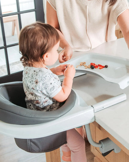 Child using a portable dining chair at a kitchen counter with a woman's assistance.