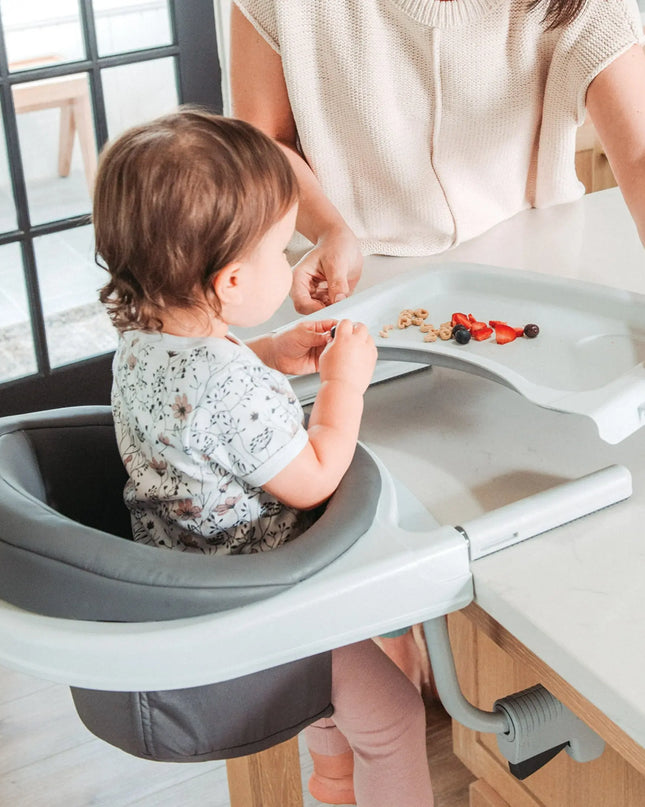 Child using a portable dining chair at a kitchen counter with a woman's assistance.