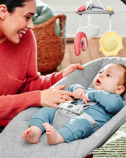 Woman interacting with a baby in a gray bouncer with colorful mobile.