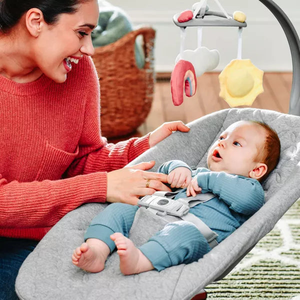 Woman interacting with a baby in a gray bouncer with colorful mobile.