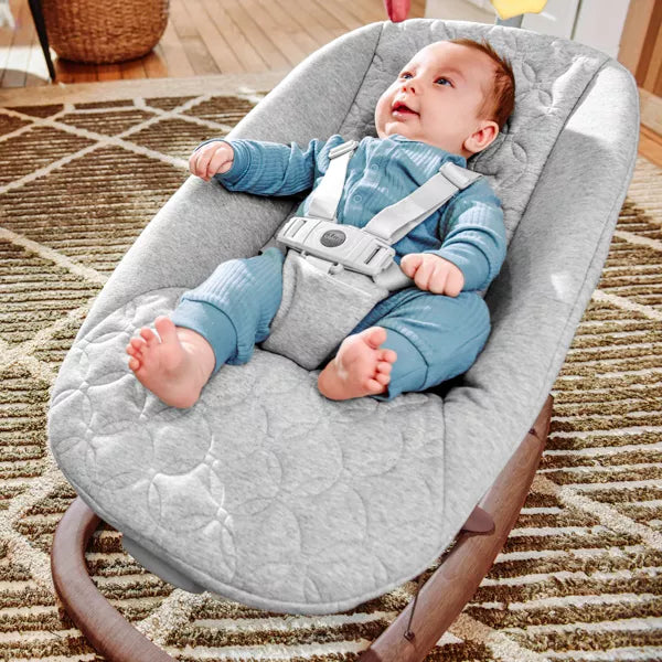 Baby in a gray baby bouncer on a carpeted floor
