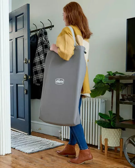 Woman carrying a gray baby carrier with ' Chicco' branding into a room.
