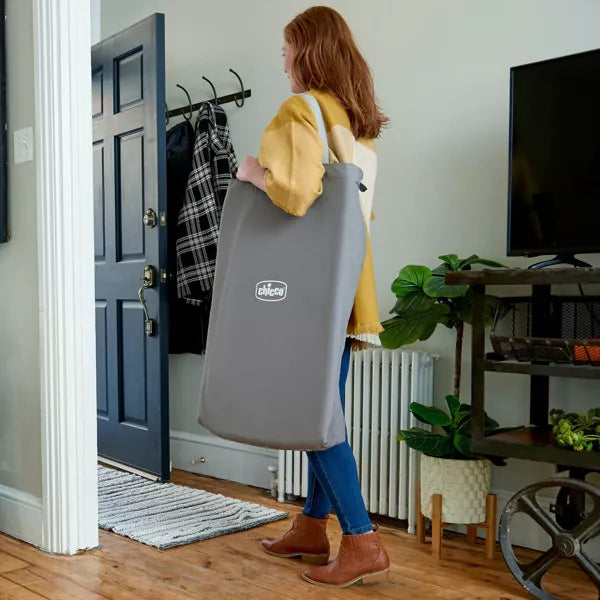 Woman carrying a gray baby carrier with ' Chicco' branding into a room.