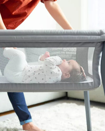 Baby lying in a gray mesh crib with a person holding the crib's handle.
