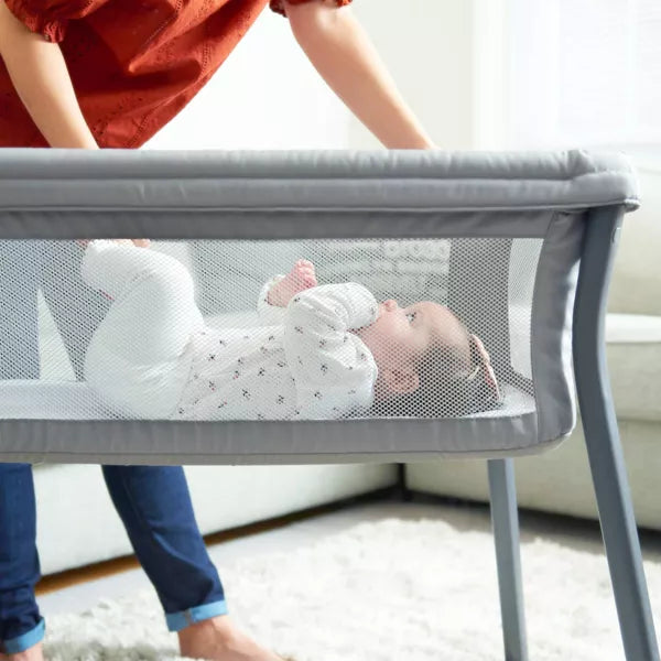 Baby lying in a gray mesh crib with a person holding the crib's handle.