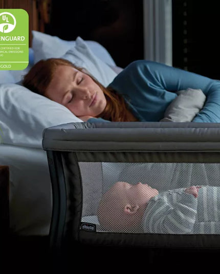 Woman lying in bed with a baby in a crib, both sleeping, with a Greenguard Gold label in the corner.