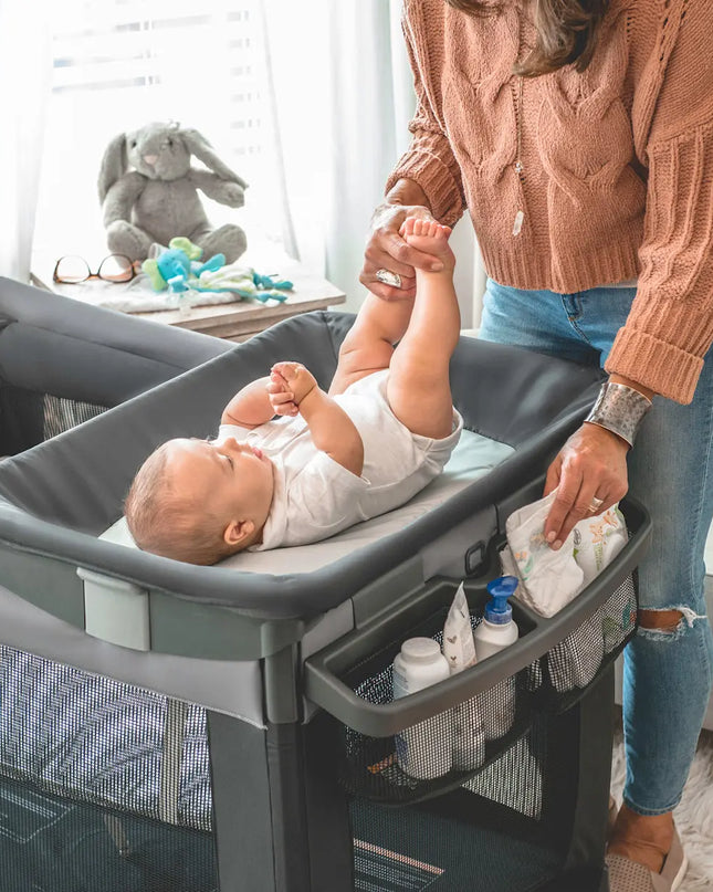 Baby lying in a portable crib with a woman standing beside it, surrounded by toys and baby items.