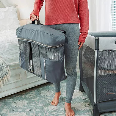 Person holding a gray travel crib bag in a bedroom setting