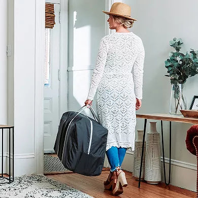 Woman in a white lace dress and straw hat holding a black suitcase in a room with a table and plant.