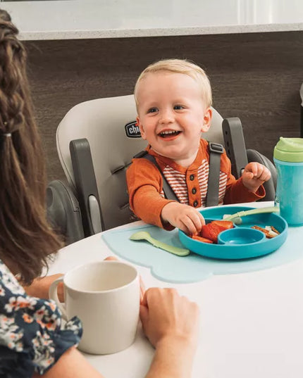 Child in a high chair with a tray of food, smiling, with an adult sitting next to them.