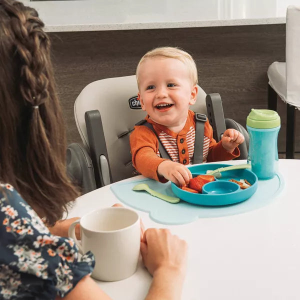 Child in a high chair with a tray of food, smiling, with an adult sitting next to them.