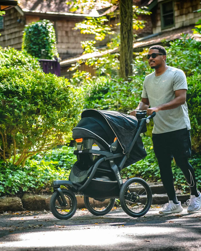 Man pushing a stroller in a park-like setting with greenery and a house in the background.