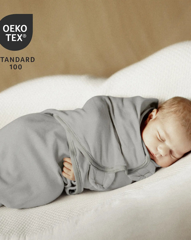 Newborn baby swaddled in gray cloth on a white blanket with OEKO-TEX Standard 100 logo.
