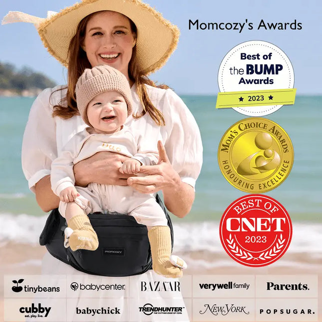 Woman holding a baby in a carrier on a beach with awards displayed.