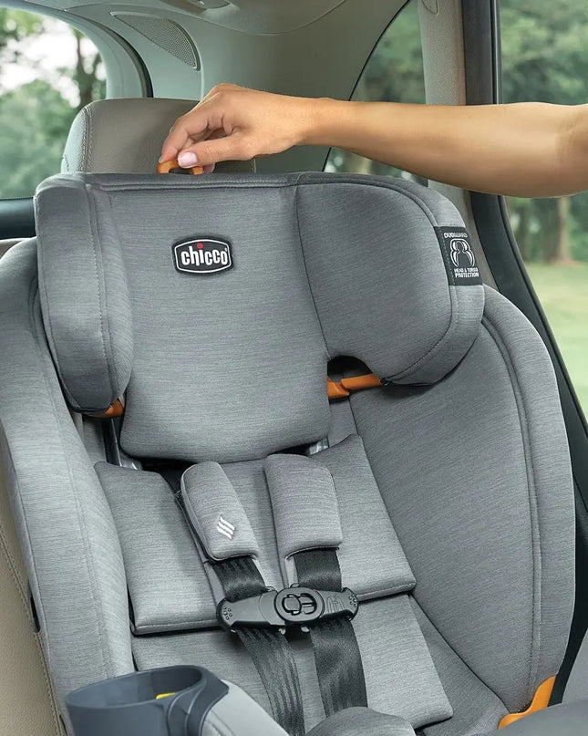 Gray Chicco car seat in a vehicle with a hand adjusting it, surrounded by greenery outside.