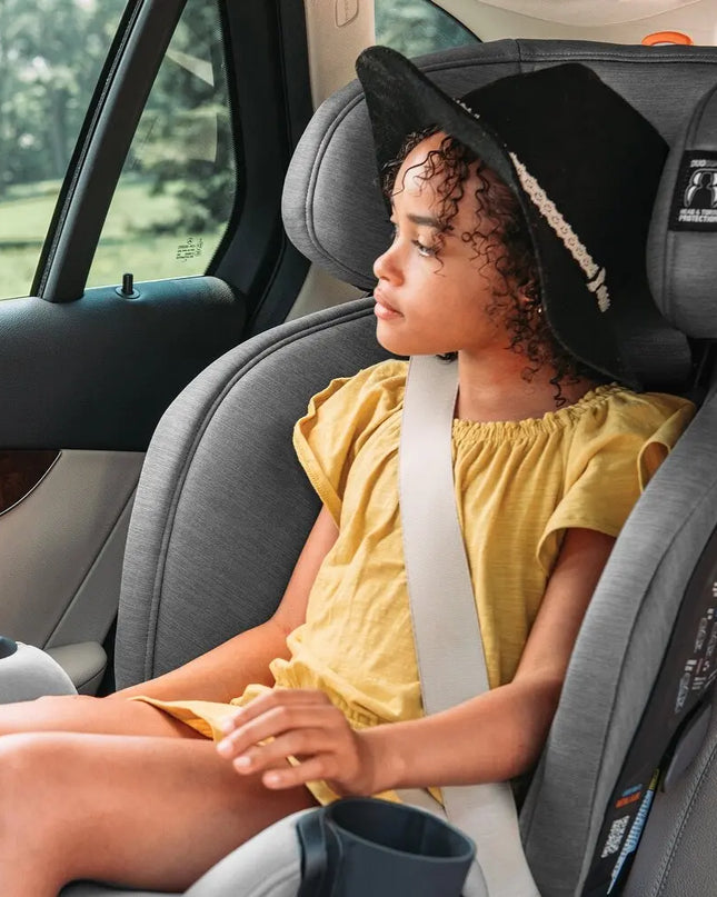 Child in a car seat wearing a yellow shirt and black hat, with a blurred outdoor background